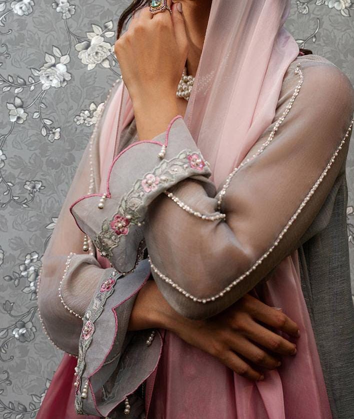 A woman wearing a pink and gray dress with intricate embroidery and a matching pink shawl, standing against a gray floral background.