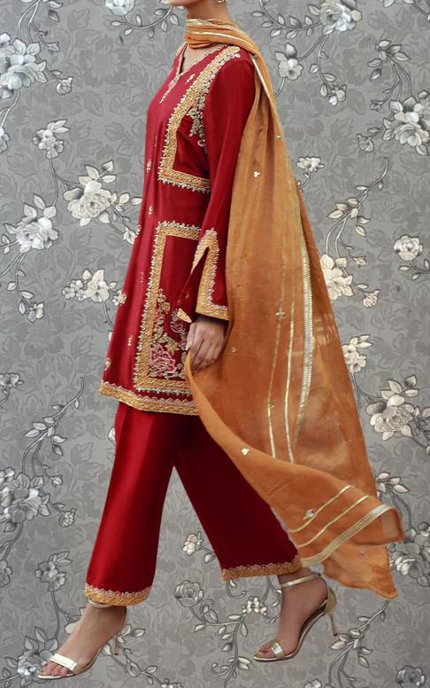 A person wearing a red and gold traditional outfit with a matching gold and gold shawl, standing against a gray floral background.
