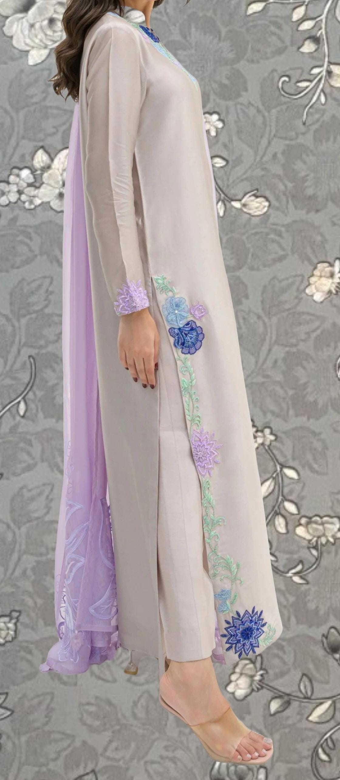 A woman wearing a long, light-colored dress with floral embroidery on the side, standing against a gray floral background.