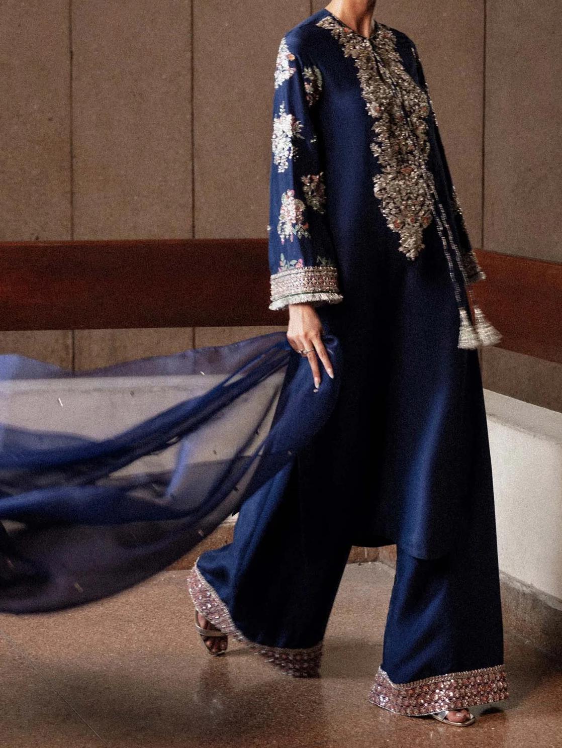 A person wearing a long navy blue dress with intricate embroidery and a flowing blue fabric draped over their body.