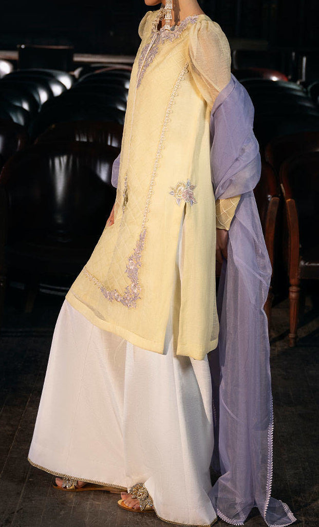 A person wearing a yellow and white dress with a purple shawl is standing in a dimly lit room with wooden chairs in the background.