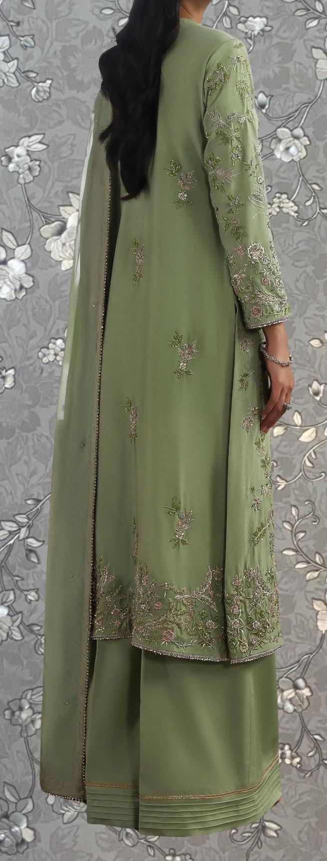 a person wearing a green dress with intricate embroidery, standing against a gray floral background.