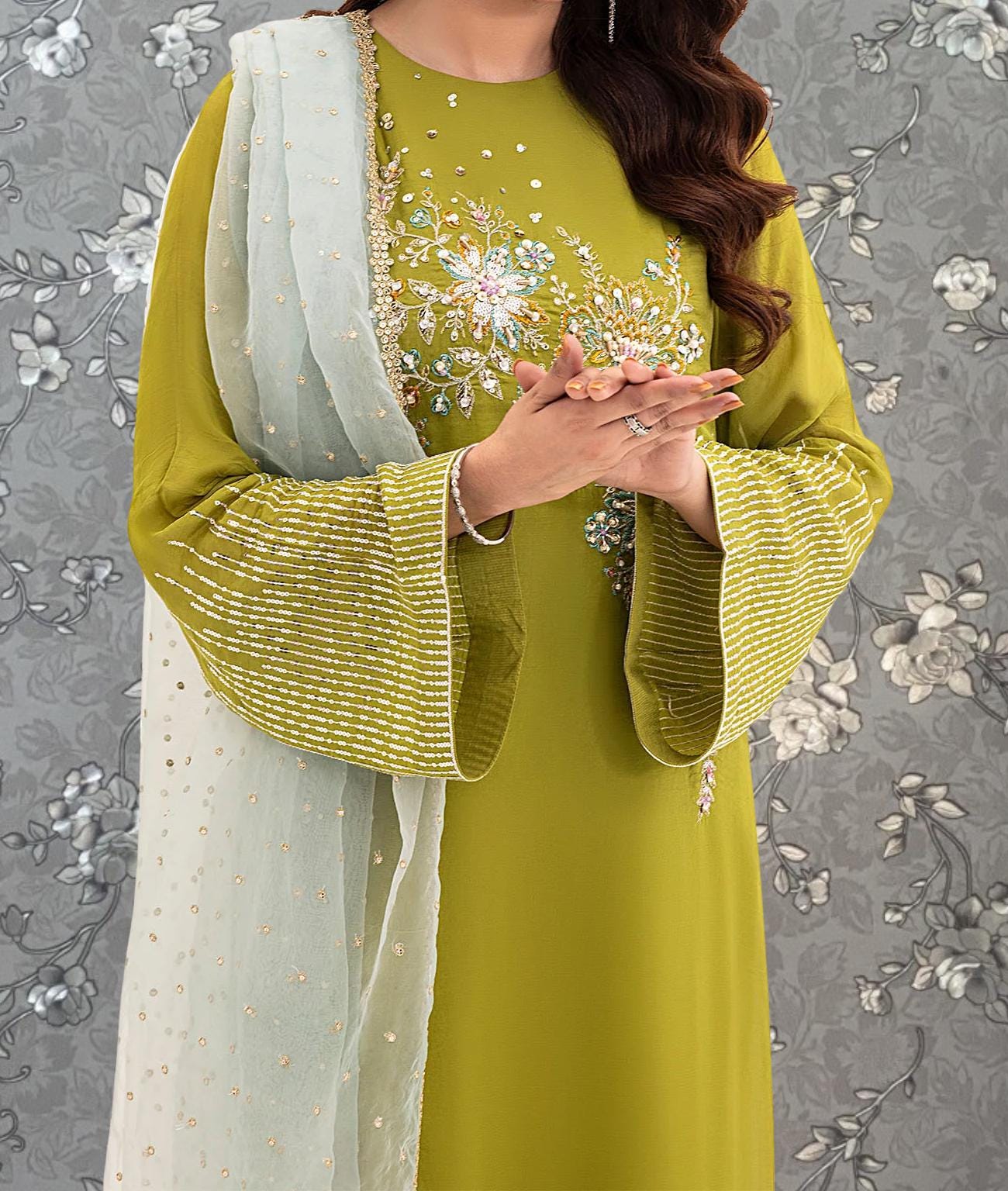 A woman wearing a green dress with gold embroidery and a white shawl stands against a gray floral background.