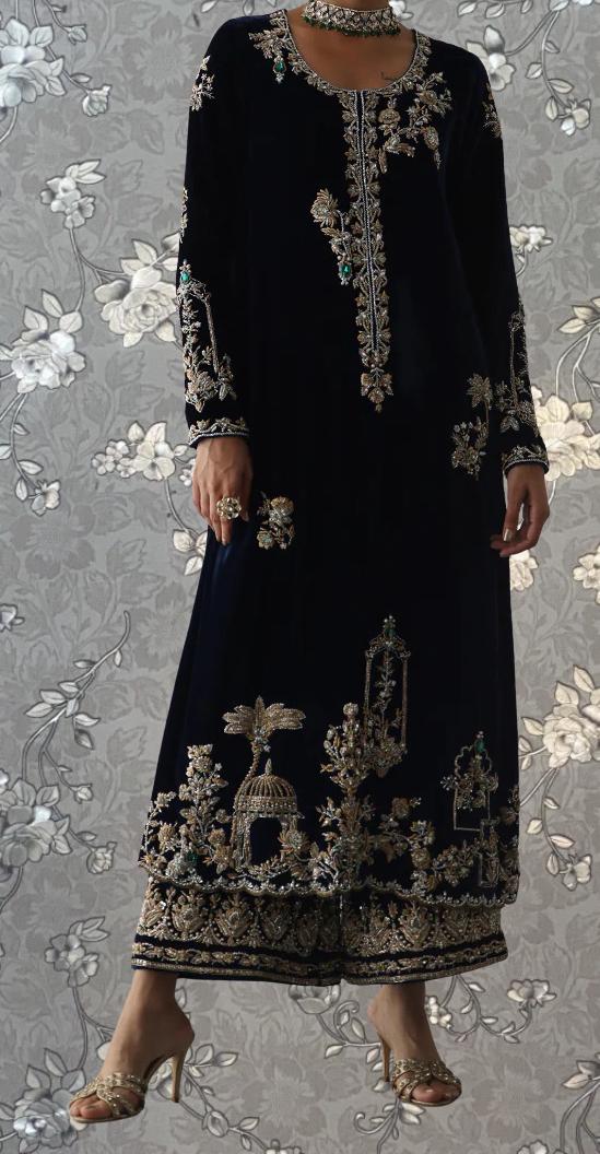 A woman wearing a long, black dress with intricate gold embroidery and floral patterns, standing against a gray floral background.
