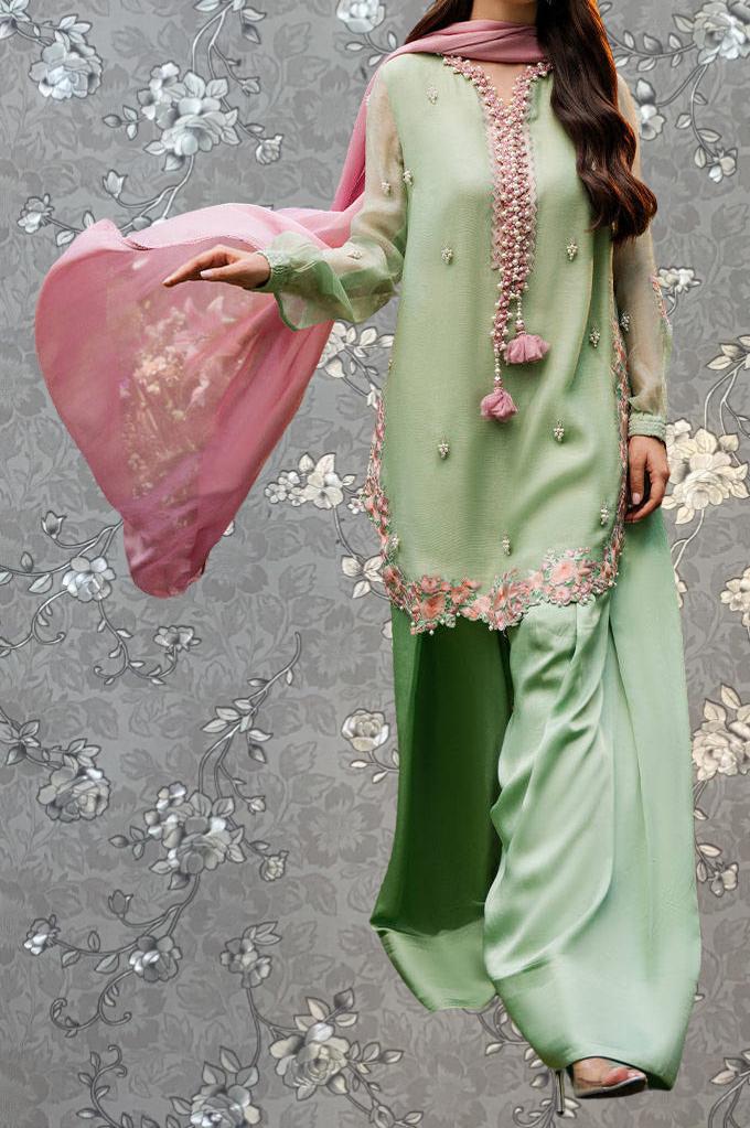 A woman wearing a green and pink dress with floral embroidery and a pink shawl is standing against a gray floral background.