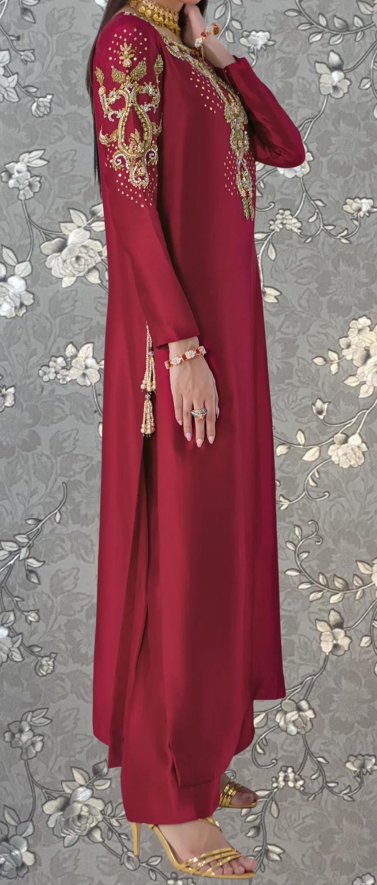 A woman wearing a long, red dress with gold embroidery and tassels, standing against a gray floral background.