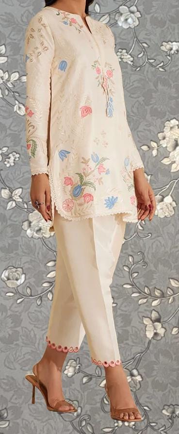 A woman wearing a white dress with floral embroidery and high heels is standing against a gray floral background.