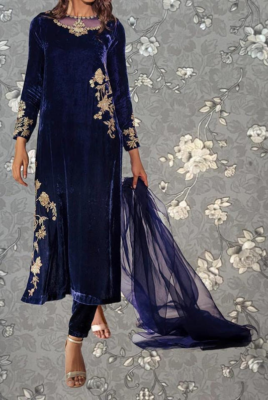 A woman wearing a long, blue velvet dress with gold embroidery stands against a gray floral background.