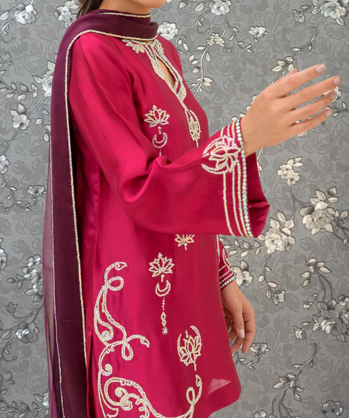 A woman wearing a vibrant red dress with intricate embroidered designs, including floral patterns and a crescent moon motif, stands against a gray floral background.