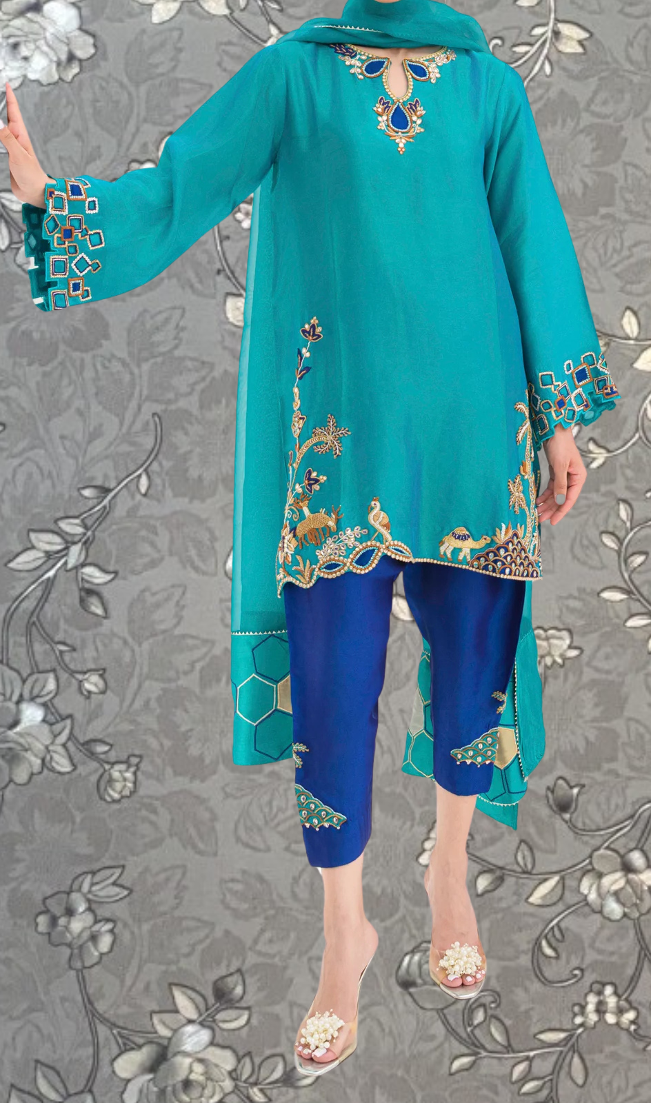 A person wearing a turquoise dress with intricate embroidery and blue pants, standing against a gray floral background.