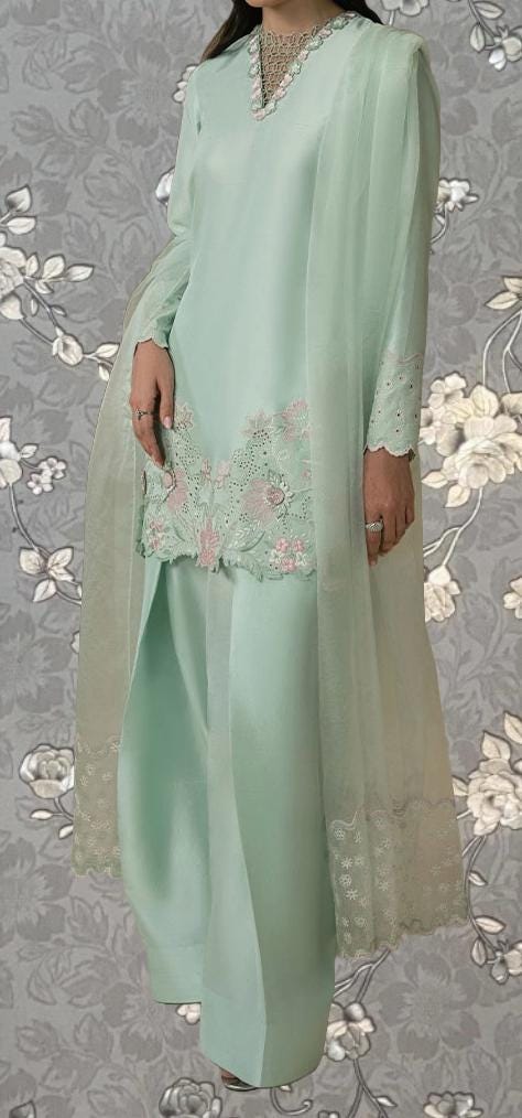 A woman wearing a mint green dress with intricate embroidery and lace trim, standing against a gray floral background.