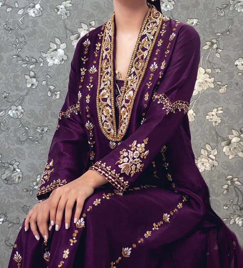 A woman wearing a purple and gold dress with intricate embroidery and patterns, sitting against a gray floral background.
