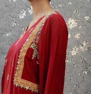 A woman wearing a red dress with gold embroidery stands against a gray floral background.