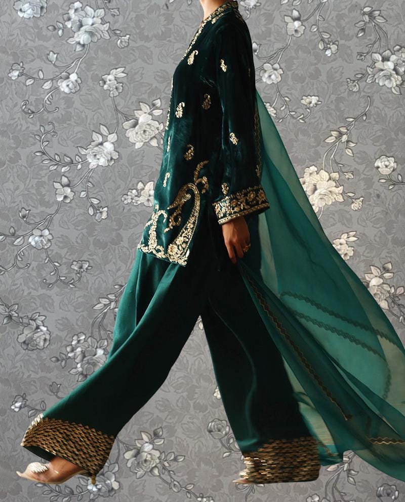 A person wearing a green velvet dress with gold embroidery and a long flowing green shawl, walking against a gray floral background.