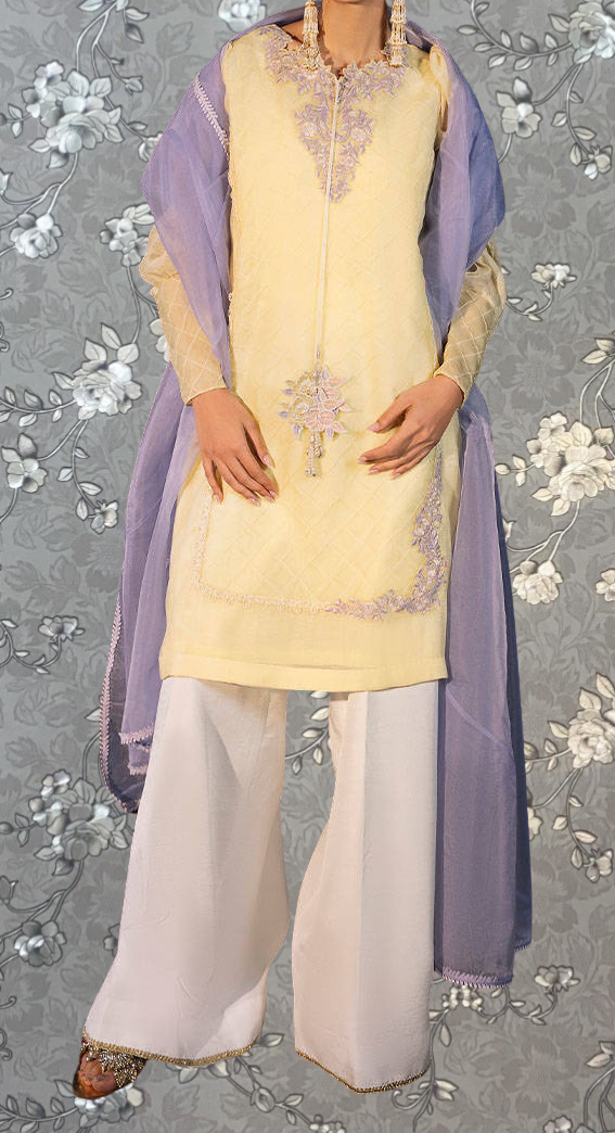 A person wearing a yellow and purple dress with a matching purple shawl, standing against a gray floral background.