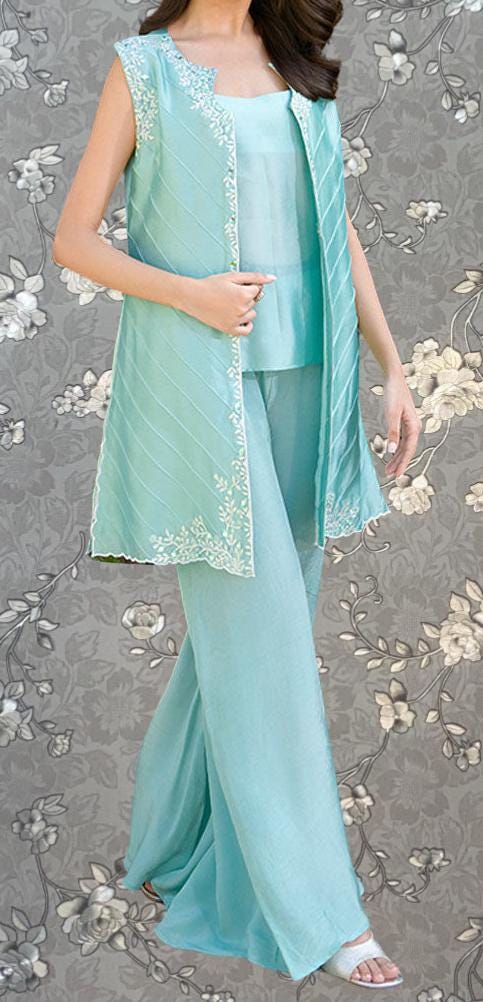 A woman wearing a mint green sleeveless top and matching pants, standing against a gray floral background.