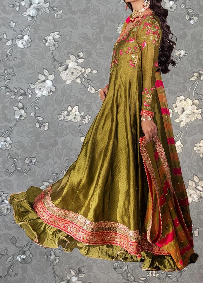 A woman wearing a long, green dress with intricate embroidery and a matching skirt, standing against a gray floral background.