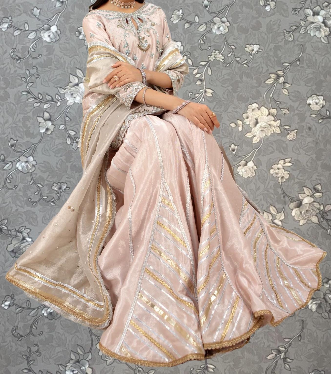 A woman wearing a pink and gold dress with intricate embroidery and gold trim, sitting against a gray floral background.