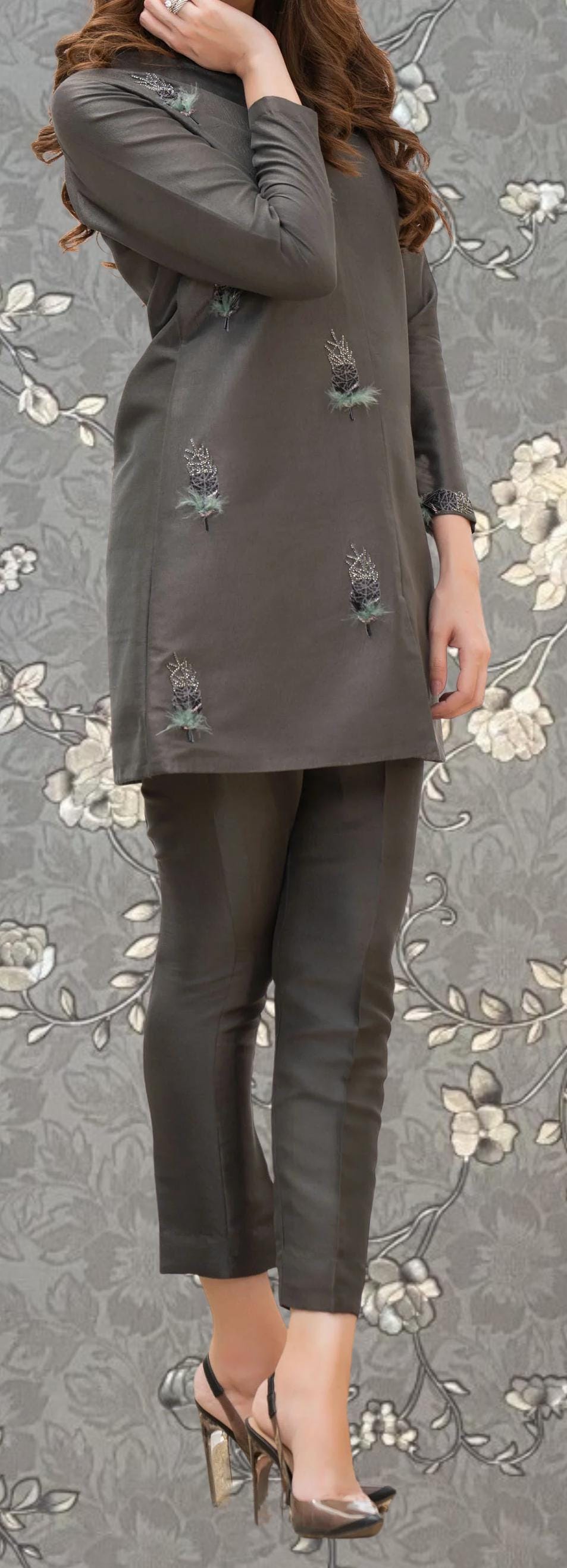 A woman wearing a gray top with floral embroidery and black pants stands against a gray floral background.