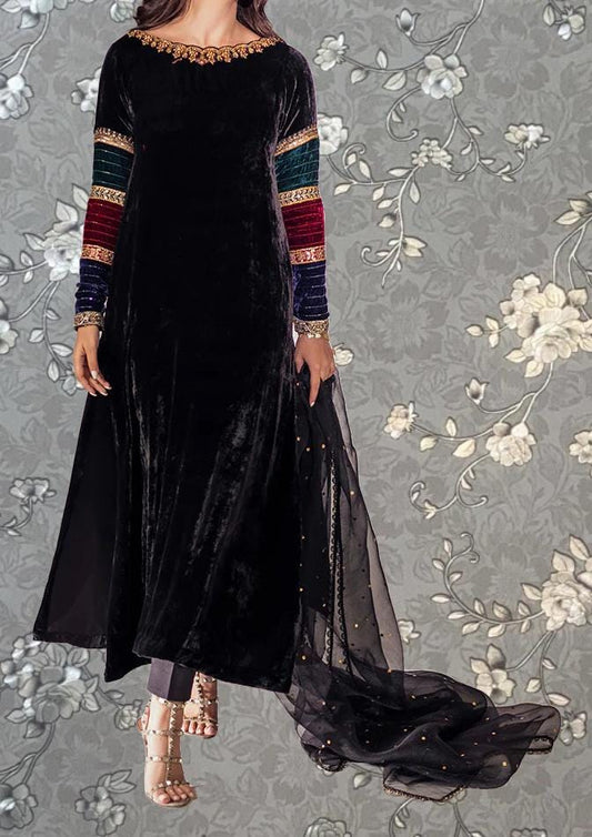 A woman wearing a long black velvet dress with colorful sleeves and a sheer black skirt, standing against a gray floral background.