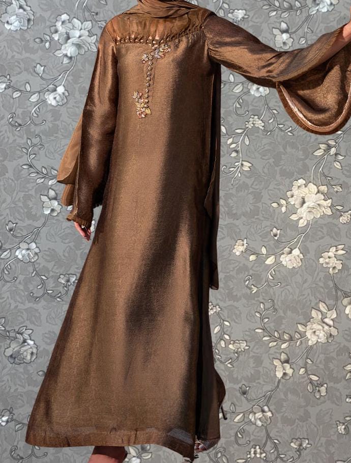 a person wearing a long, brown dress with a floral pattern on the back. The dress has a long, flowing silhouette and appears to be made of a shiny, metallic fabric. The person is standing against a gray floral background, which complements the overall design of the dress.