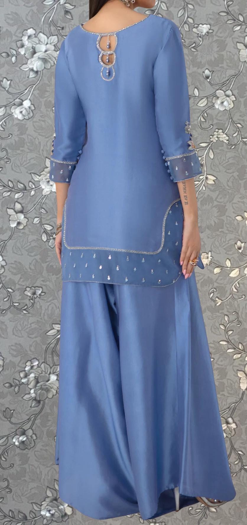 A woman wearing a blue dress with silver embellishments and a floral pattern on the background.