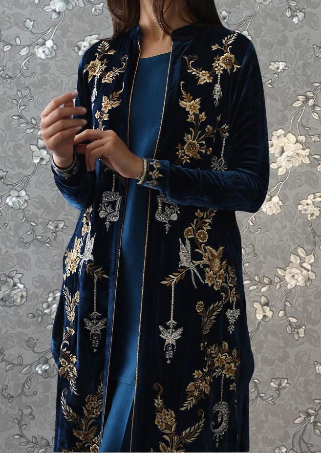 A woman wearing a long, blue velvet coat with gold embroidery stands against a gray floral background.