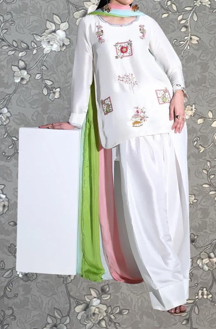 A woman wearing a white dress with colorful embroidered designs, standing against a gray floral background.