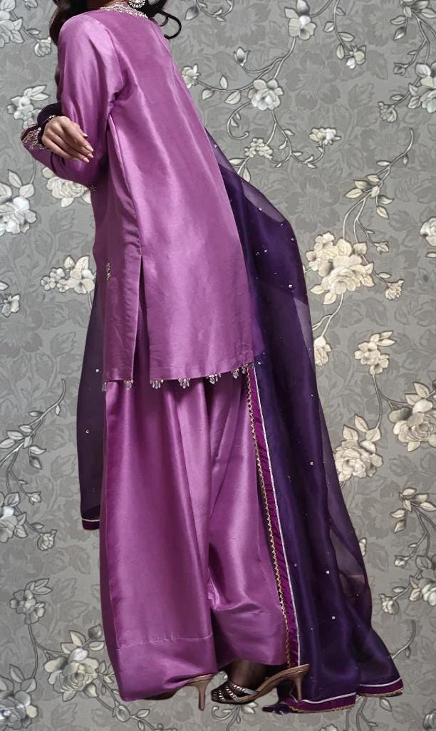 A person wearing a long purple dress with a purple shawl draped over it, standing against a gray floral background.