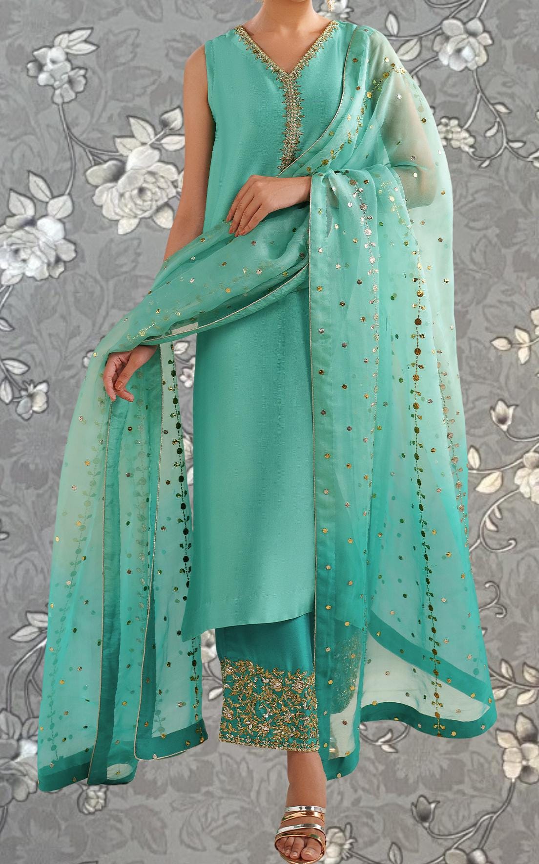 A woman wearing a turquoise dress with a matching turquoise shawl and gold embellishments, standing against a gray floral background.