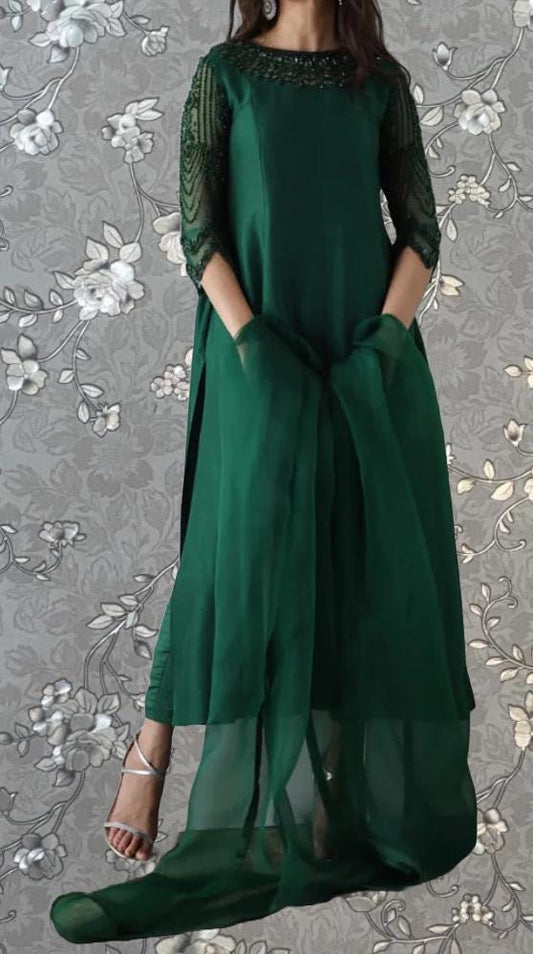 A woman wearing a long, green dress with intricate beading and sleeves stands against a gray floral background.