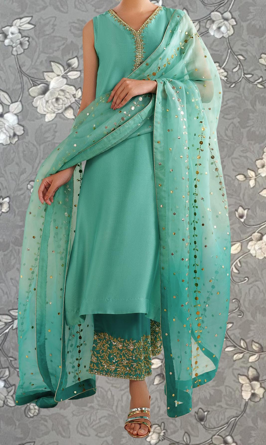A woman wearing a turquoise dress with gold embellishments and a matching turquoise shawl, standing against a gray floral background.