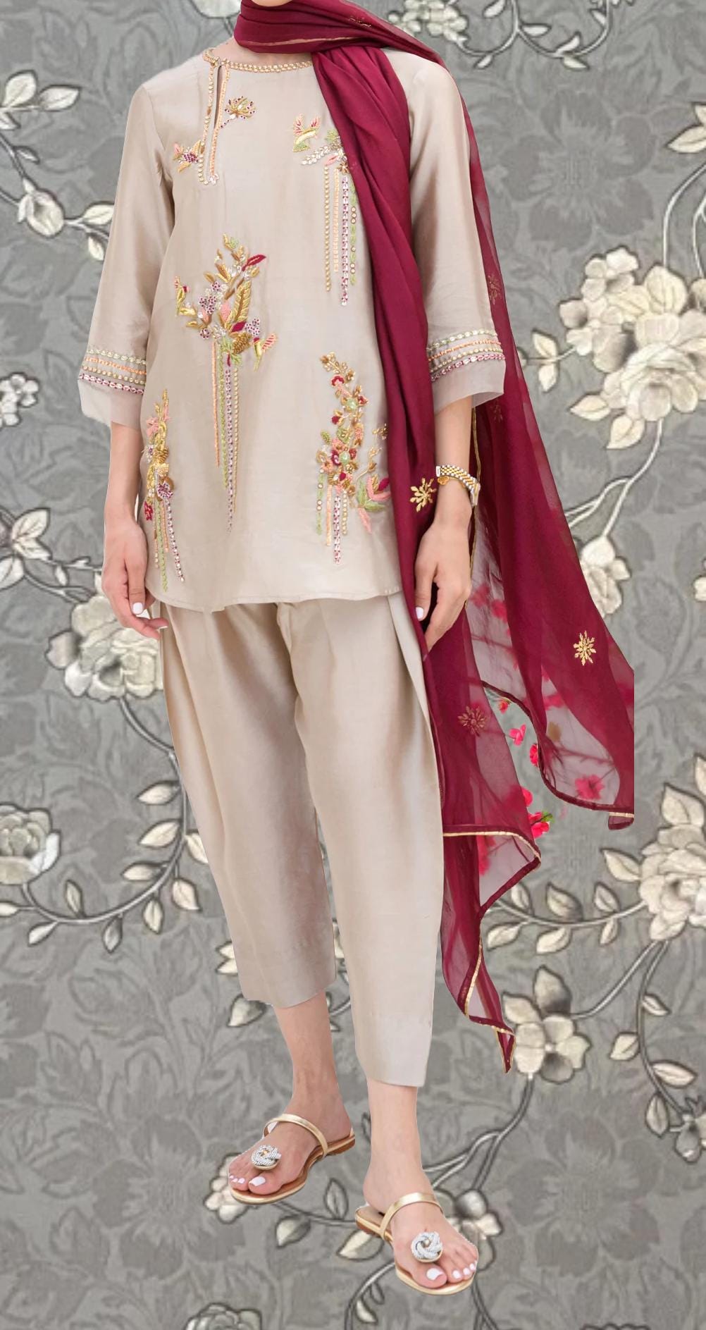 A person wearing a beige dress with intricate embroidery and a red scarf, standing against a gray floral background.