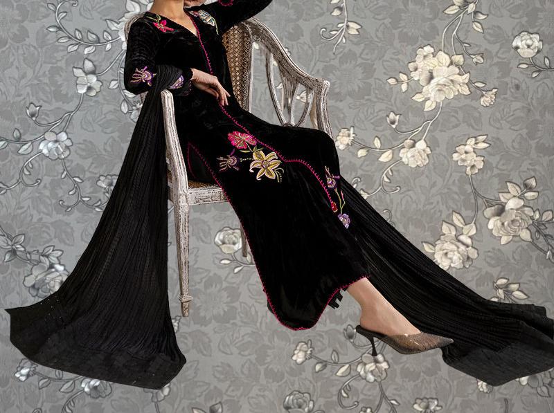 A woman wearing a long black dress with floral embroidery is sitting on a chair against a gray floral wallpaper background.
