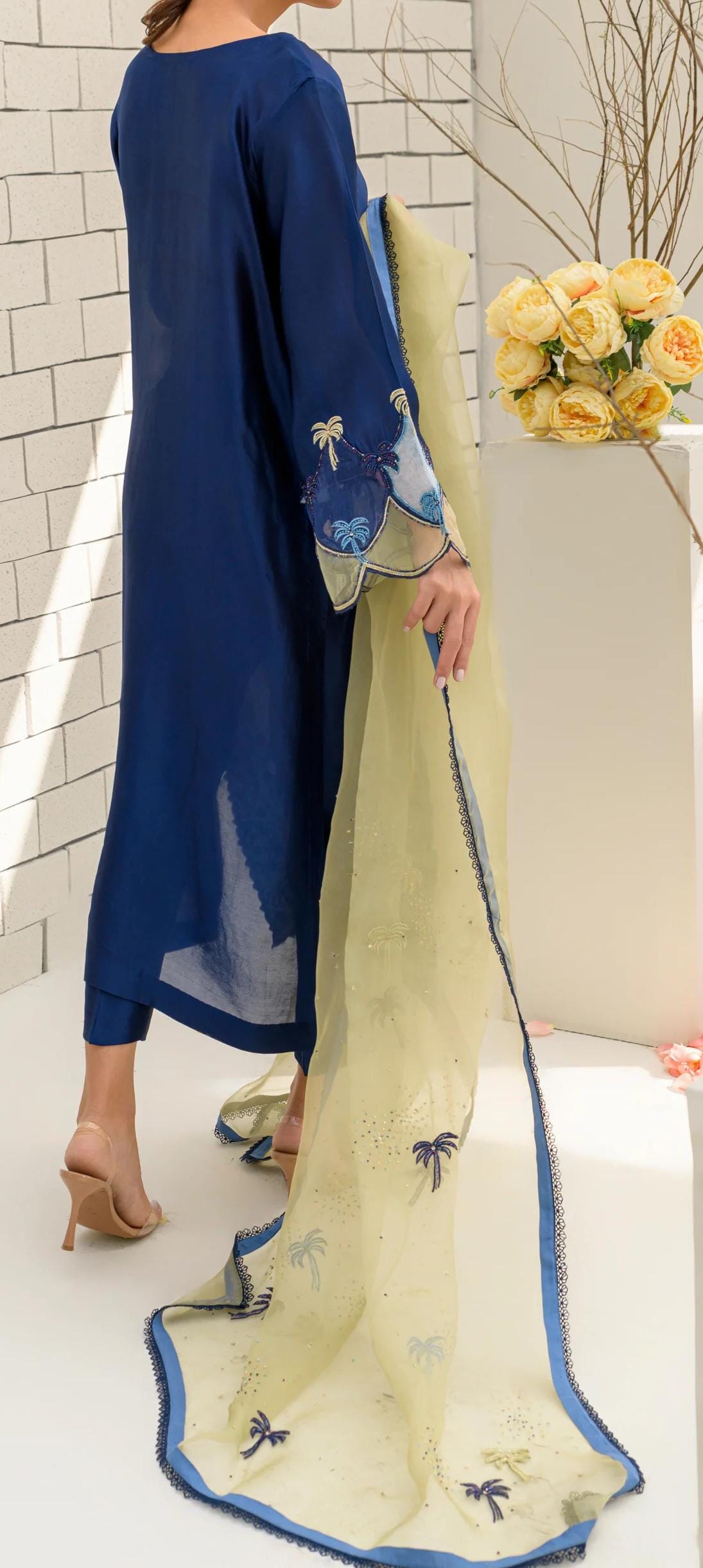 A person wearing a blue dress with yellow and blue embroidered details, standing in front of a white wall with a vase of yellow flowers.