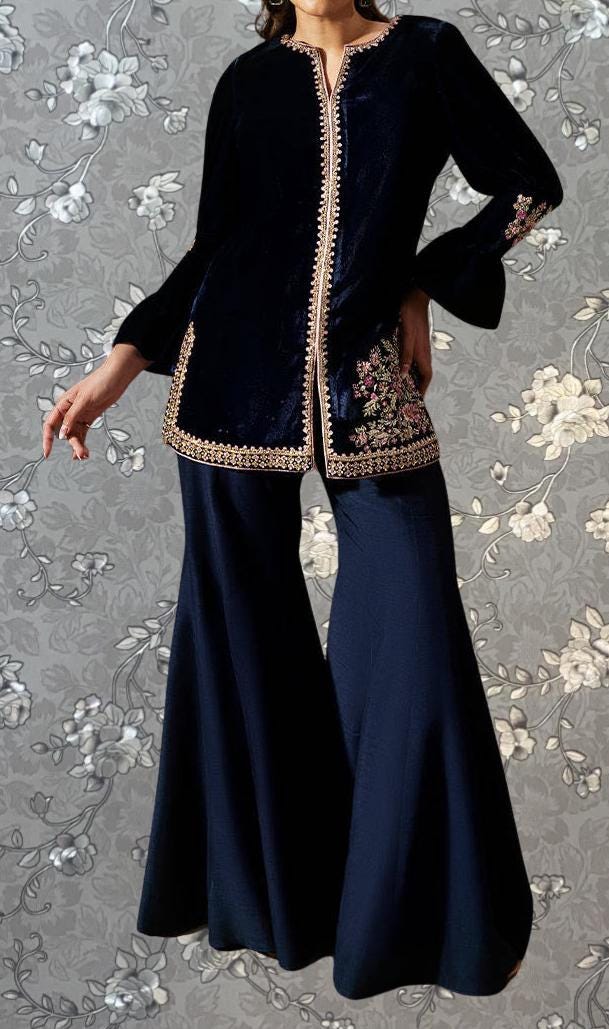 A woman wearing a navy blue velvet jacket with gold trim and matching pants stands against a gray floral background.
