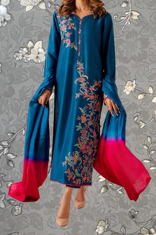 A woman wearing a long blue dress with floral embroidery and a matching red shawl is shown against a gray floral background.