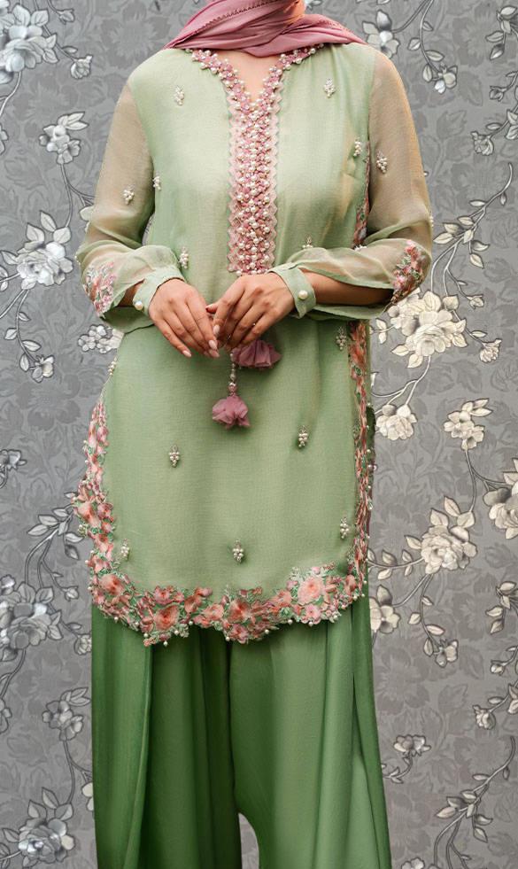 A woman wearing a green dress with pink floral accents and a matching green skirt, standing against a gray floral background.