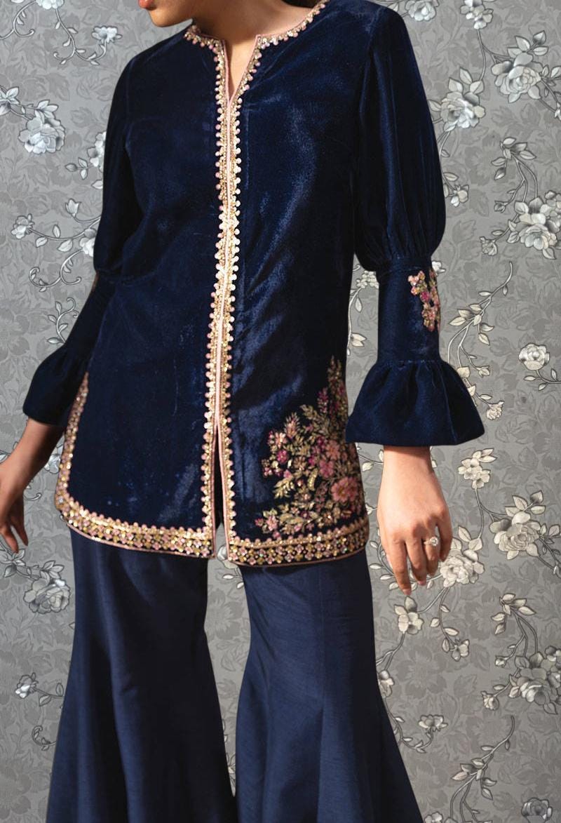 A woman wearing a navy blue velvet jacket with gold trim and floral embroidery on the sleeves, paired with matching navy blue pants.