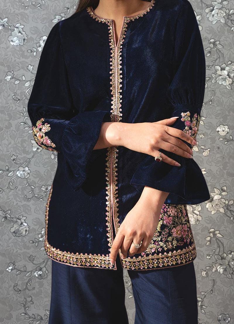 A woman wearing a navy blue velvet blouse with gold embroidery and a matching navy blue skirt, posing with her arms crossed against a gray floral background.
