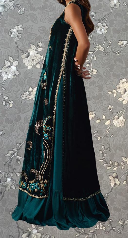 A woman wearing a long, green velvet dress with intricate gold embroidery stands against a gray floral background.