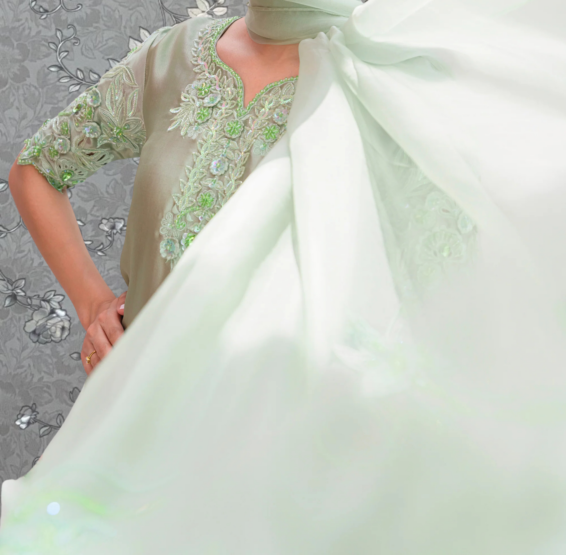 A person wearing a green and white dress with intricate embroidery stands against a gray floral background.