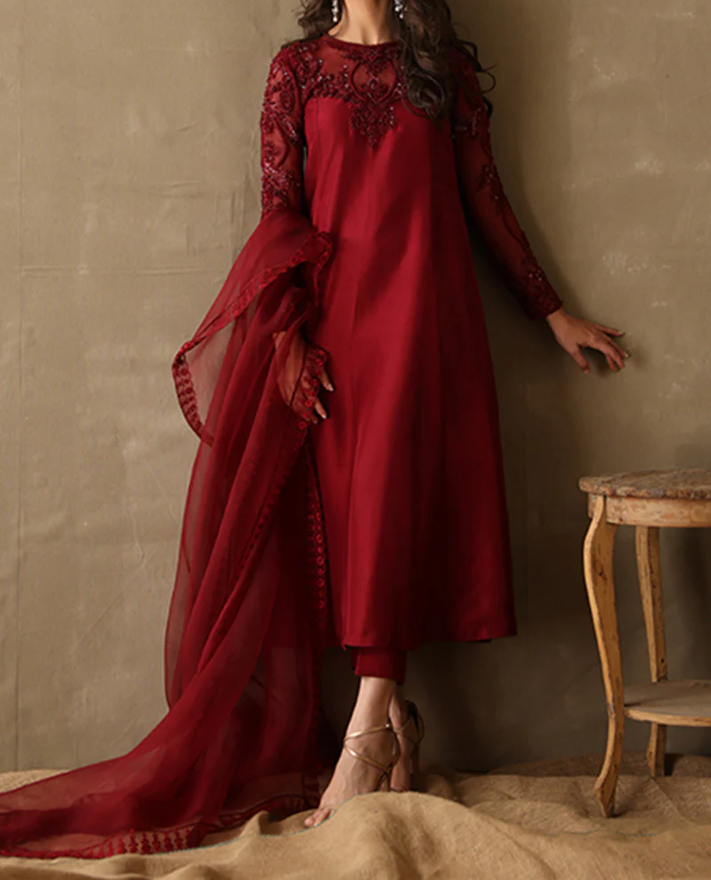 A woman wearing a long red dress with intricate embroidery stands in front of a wooden table.