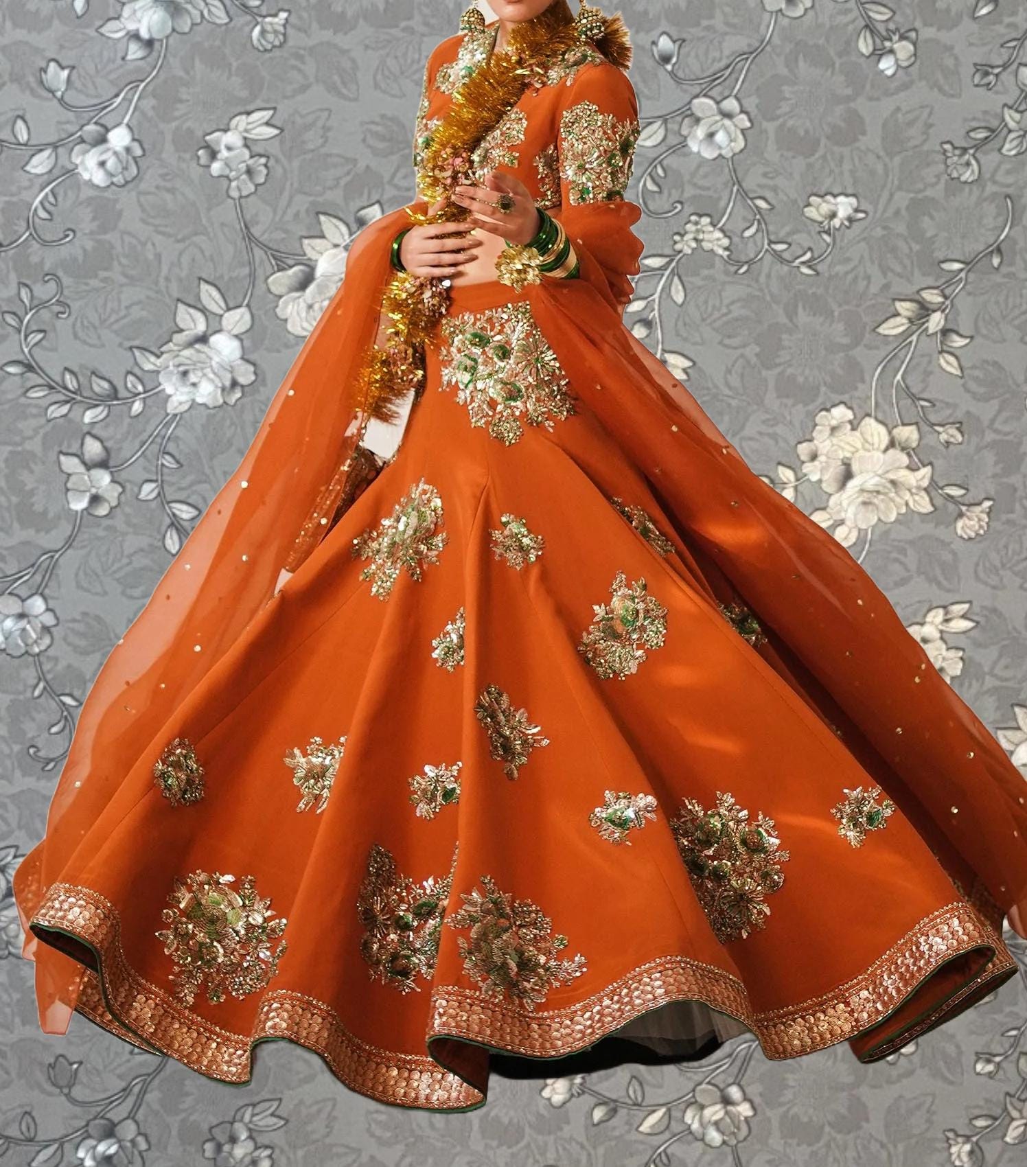 A woman wearing an ornate, orange and gold dress with intricate embroidery and a flowing skirt, standing against a gray floral background.