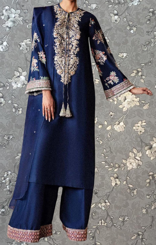 A person wearing a long navy blue dress with intricate embroidered patterns and tassels, standing against a gray floral background.