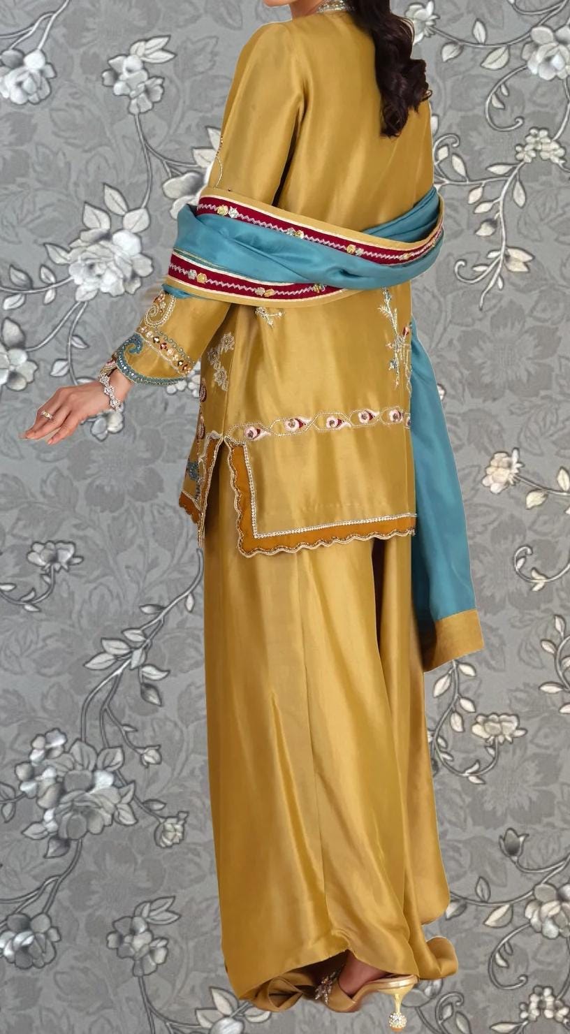 A person wearing a gold and blue dress with intricate embroidery and patterns, standing against a gray floral background.