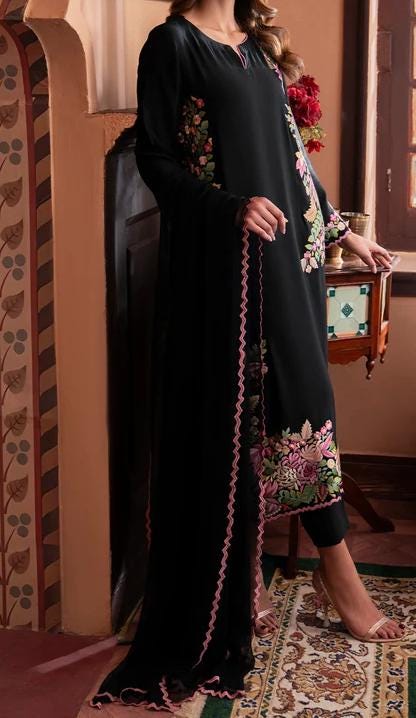 A woman wearing a long black dress with floral embroidery stands in a room with a floral patterned wall.