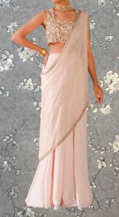 A woman wearing a pink and gold saree with a gold and beaded blouse, standing against a gray floral background.