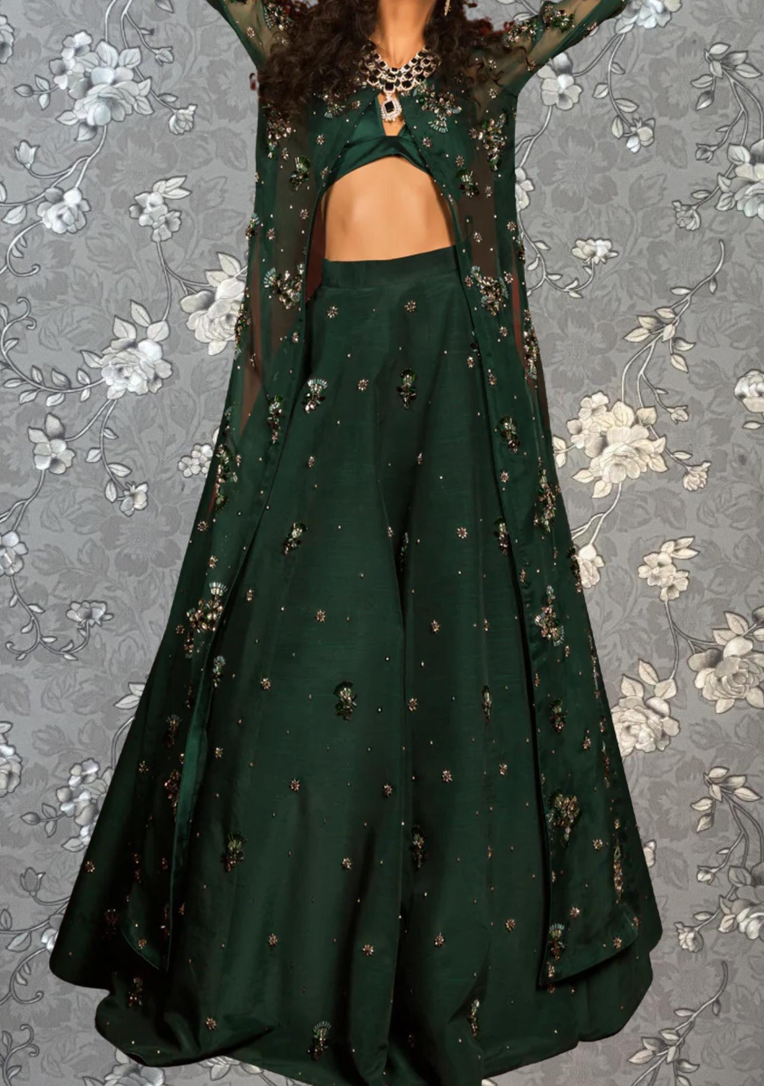 A woman wearing a green dress with a sheer overlay and a matching green skirt, standing against a gray floral background.