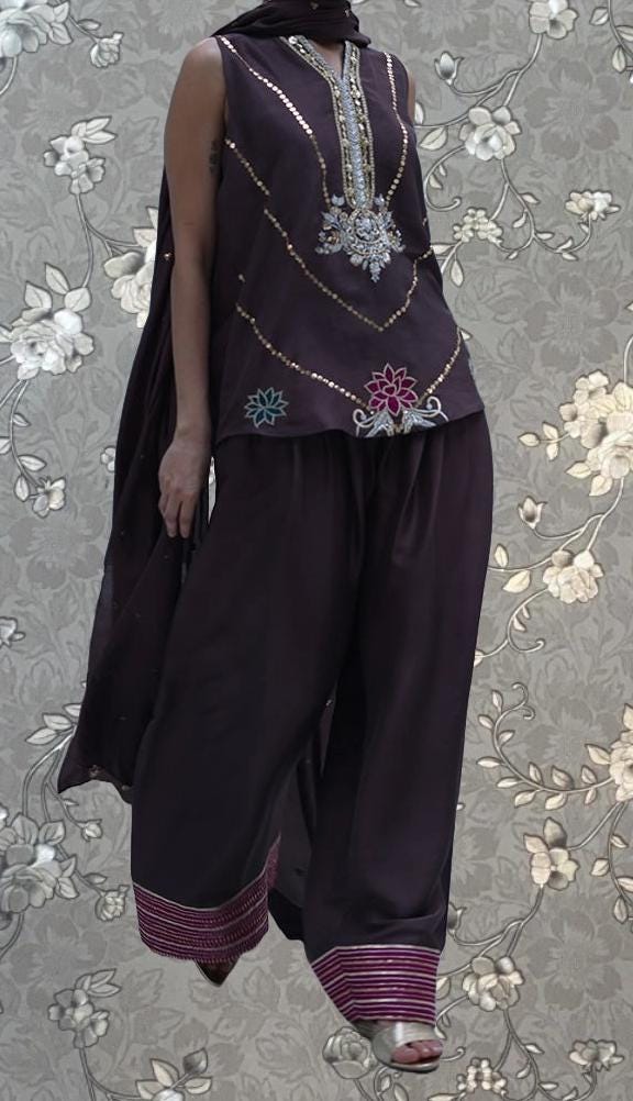 A person wearing a purple dress with intricate embroidery and floral patterns, standing against a floral background.